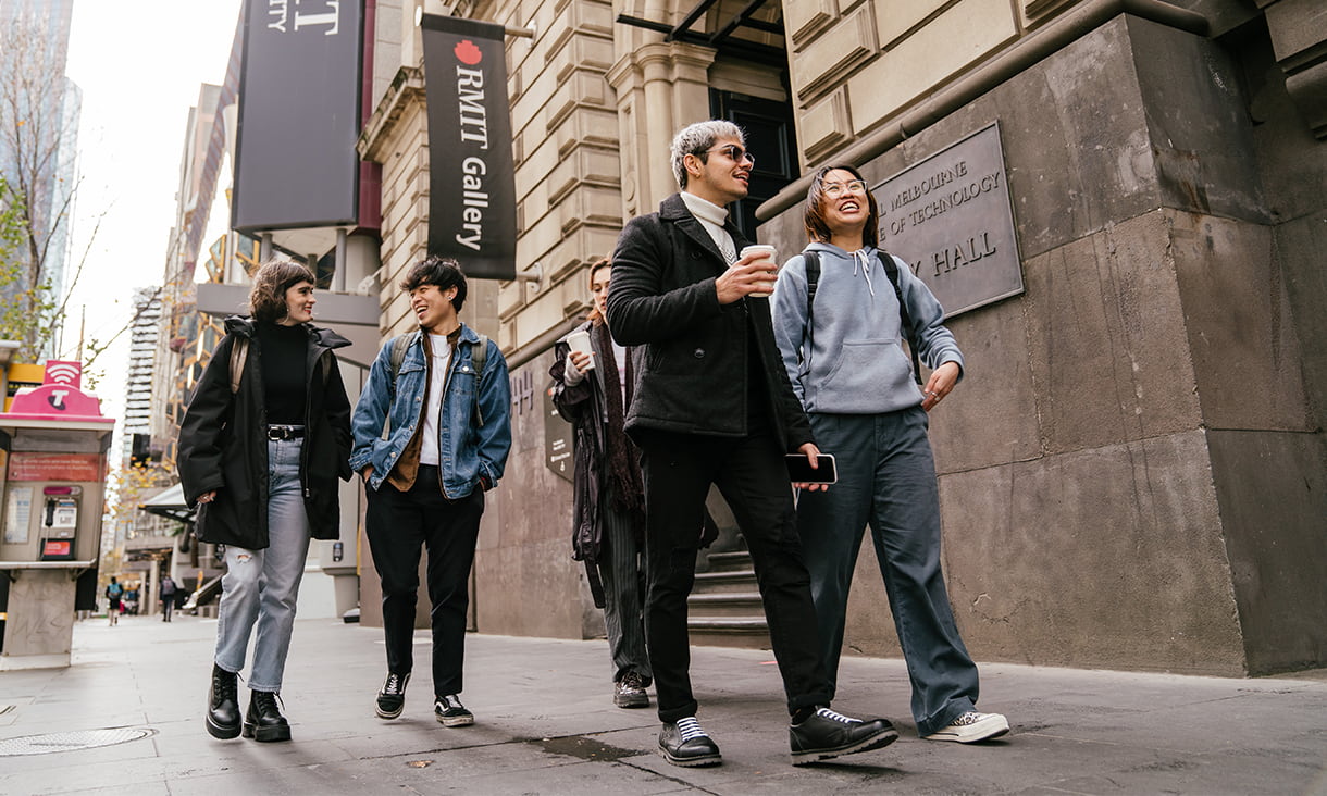 Four casually dressed students walking through Swanston street. The buildings surrounding them are brown brick and a pay-phone is visible in front of trees lining the street.